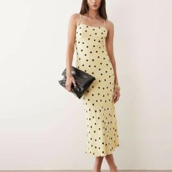 robes à pois jaune longue dos ouvert décolleté cœur caraco portée par un mannequin élégant tenant une pochette noire