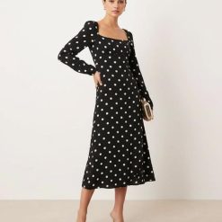 robes longues noires à pois encolure carrée petite delilah manches longues portée par un mannequin avec pochette dorée et sandales à talons