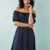 robes asymétriques à pois noués manches bouffantes plage longue bleu marine portée par une femme avec col bardot épaules dénudées sur fond bleu clair