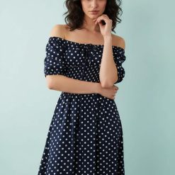 robes asymétriques à pois noués manches bouffantes plage longue bleu marine portée par une femme avec col bardot épaules dénudées sur fond bleu clair
