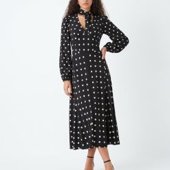 robes longues noires à pois blancs sadie col lavallière longueur mollet à manches longues et décolleté en v portée par un mannequin avec des sandales à talons noirs